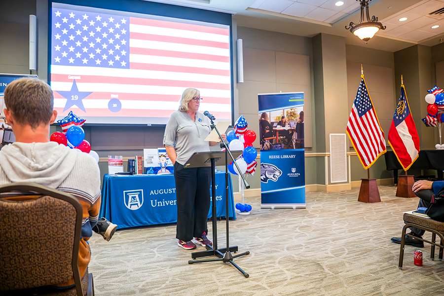 A woman stands in front of a large crowd at a microphone and reads part of the U.S. Constitution.
