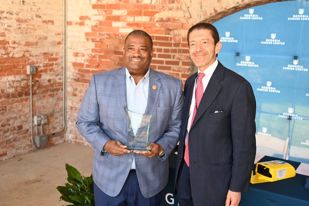 Two men smile for a photo. The man on the left is holding a glass award.