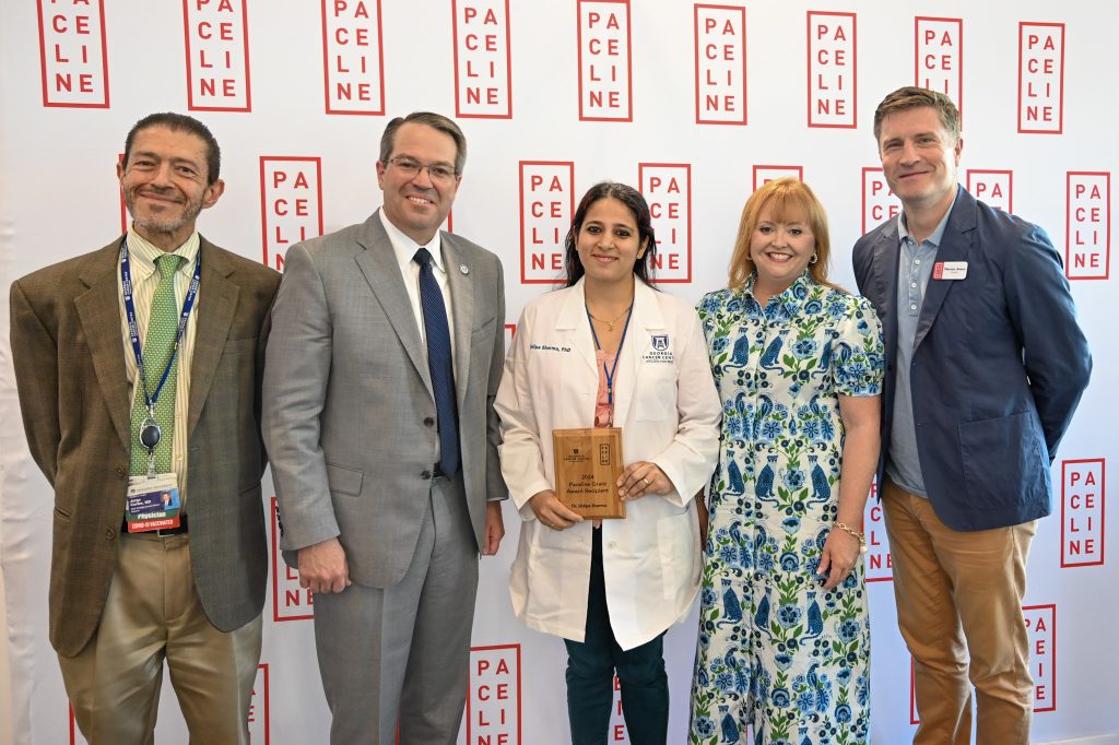 Five people, three men and two women, during an award ceremony. The woman in the middle is wearing a scientific lab coat and holding an award.