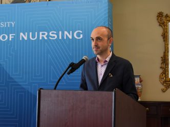A man in a suit jacket standing at a podium in front of a step and repeat that says "Nursing."