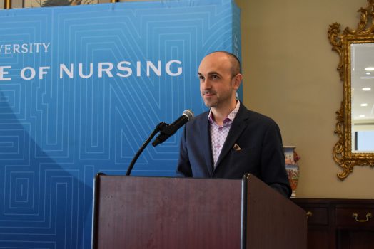 A man in a suit jacket standing at a podium in front of a step and repeat that says "Nursing."