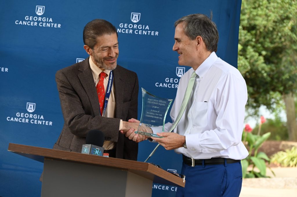Two men shaking hands at a podium with a navy Georgia Cancer Center backdrop behind them. The man on the right is holding a glass award.