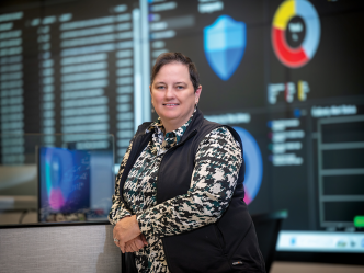 A woman stands in a security operation center for cybersecurity. One wall of the room is covered in large screens showing graphs and information.