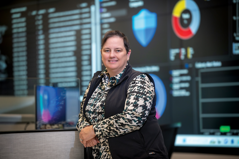 A woman stands in a security operation center for cybersecurity. One wall of the room is covered in large screens showing graphs and information.