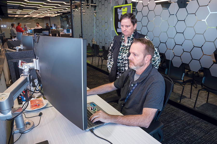 A woman looks over the shoulder of a man sitting at a computer station while he explains what he is seeing.