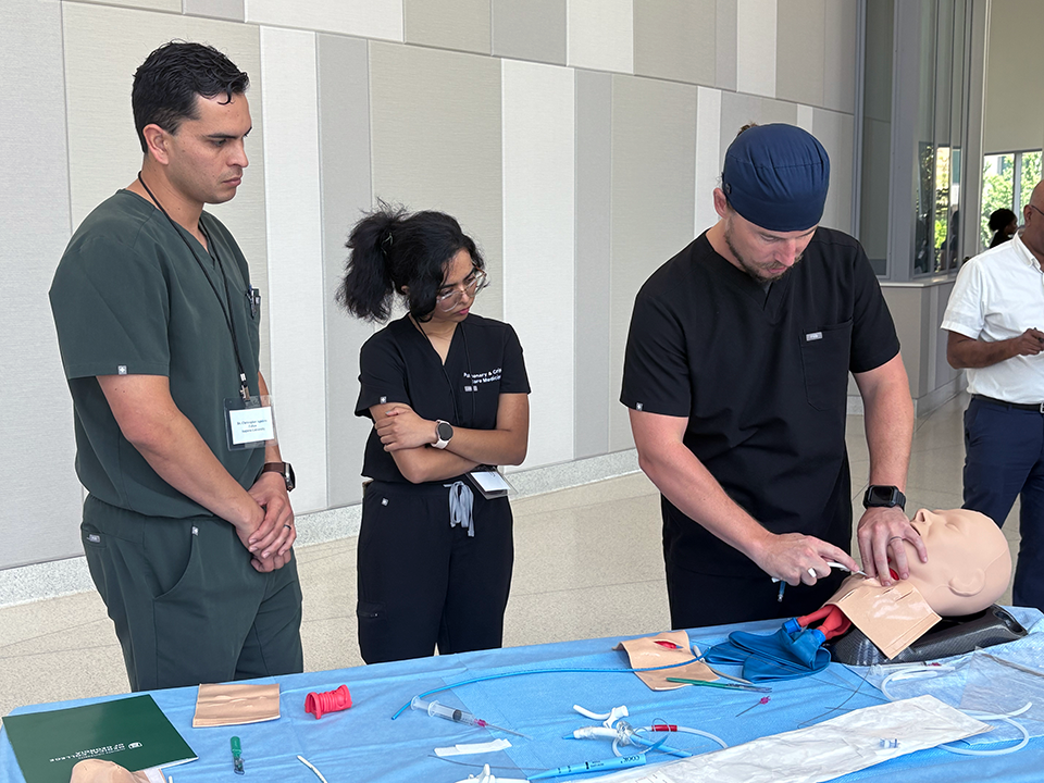 A male and female pulmonary fellow watch a pulmonologist perform a tracheostomy on a medical mannequin.