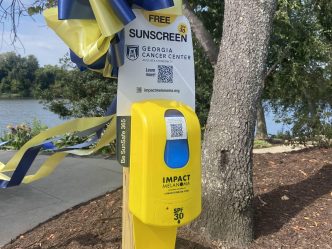 a yellow sunscreen dispenser with a yellow and blue ribbon attached to it stands next to a tree in a park. There is a sign attached to it that says "Free Sunscreen SPF 30" with a Georgia Cancer Center Augusta University logo and "Learn More" next to a QR code. At the bottom of the sign is a web address for impactmelanoma.org.