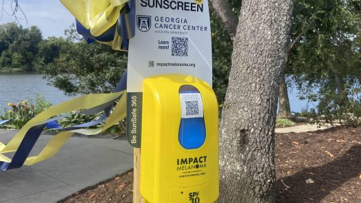 a yellow sunscreen dispenser with a yellow and blue ribbon attached to it stands next to a tree in a park. There is a sign attached to it that says "Free Sunscreen SPF 30" with a Georgia Cancer Center Augusta University logo and "Learn More" next to a QR code. At the bottom of the sign is a web address for impactmelanoma.org.