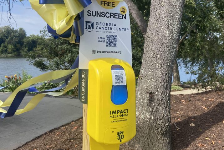 a yellow sunscreen dispenser with a yellow and blue ribbon attached to it stands next to a tree in a park. There is a sign attached to it that says "Free Sunscreen SPF 30" with a Georgia Cancer Center Augusta University logo and "Learn More" next to a QR code. At the bottom of the sign is a web address for impactmelanoma.org.