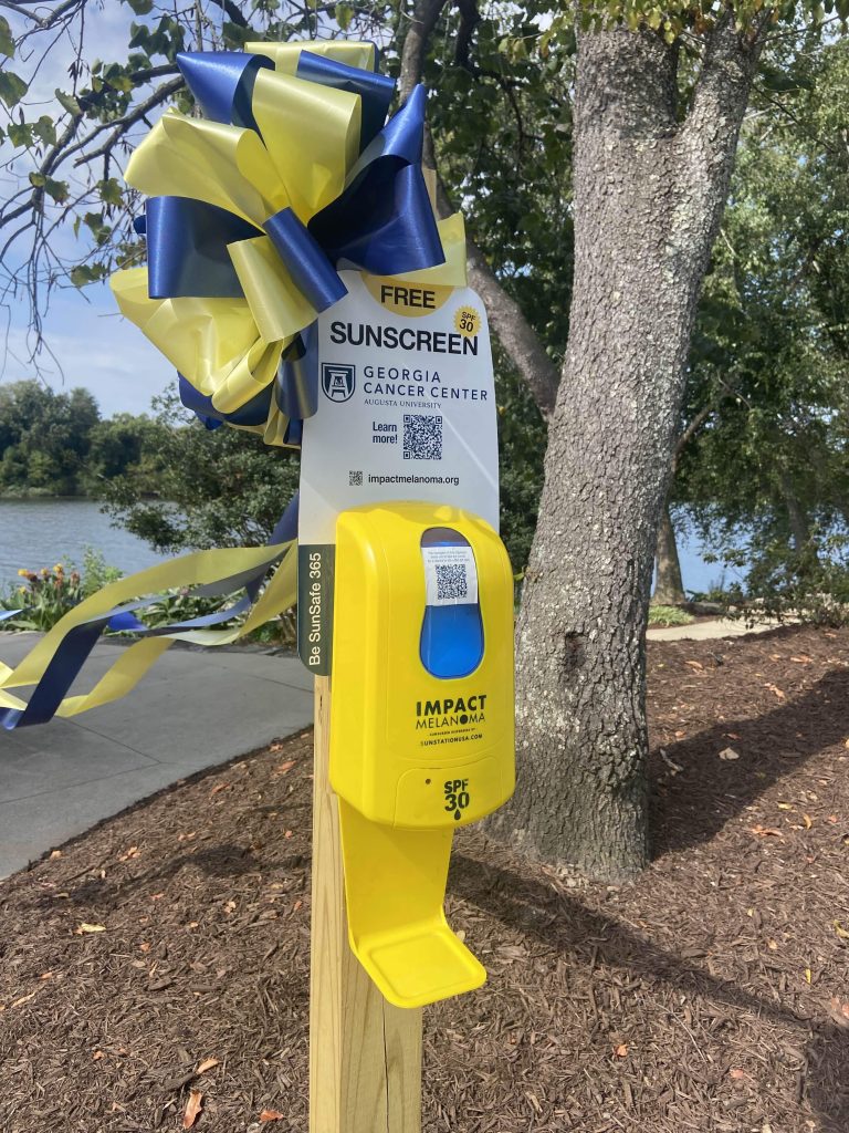a yellow sunscreen dispenser with a yellow and blue ribbon attached to it stands next to a tree in a park. There is a sign attached to it that says "Free Sunscreen SPF 30" with a Georgia Cancer Center Augusta University logo and "Learn More" next to a QR code. At the bottom of the sign is a web address for impactmelanoma.org.