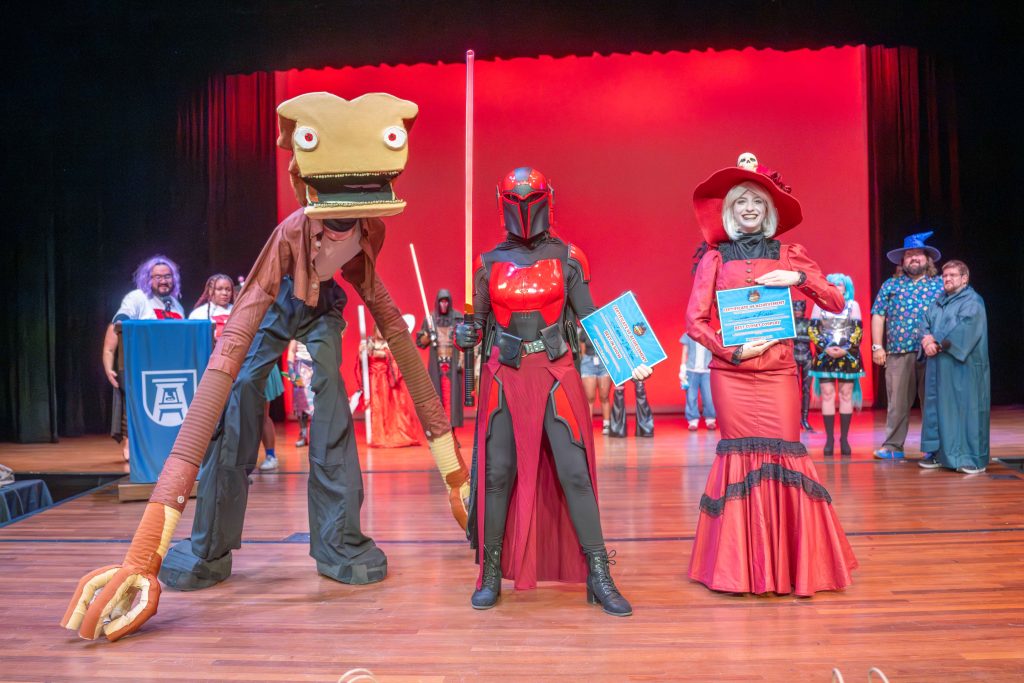 Three college-aged students in line on a stage dressed in costumes celebrate winning awards during a cosplay contest.