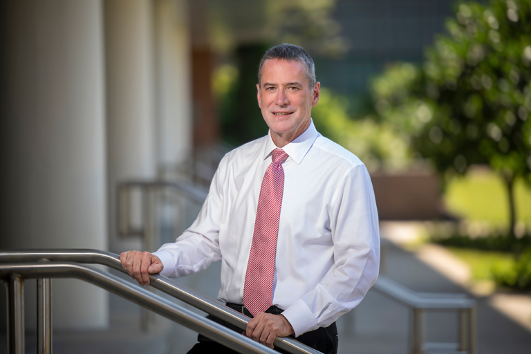 A man wearing a button down shirt and a tie stands on a set of outdoor stairs and smiles at the camera.