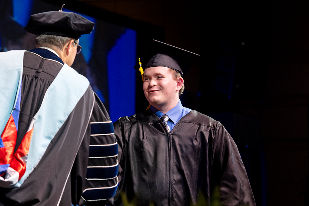 Young man in graduation cap and gown shakes the hand of another man wearing graduation regalia.