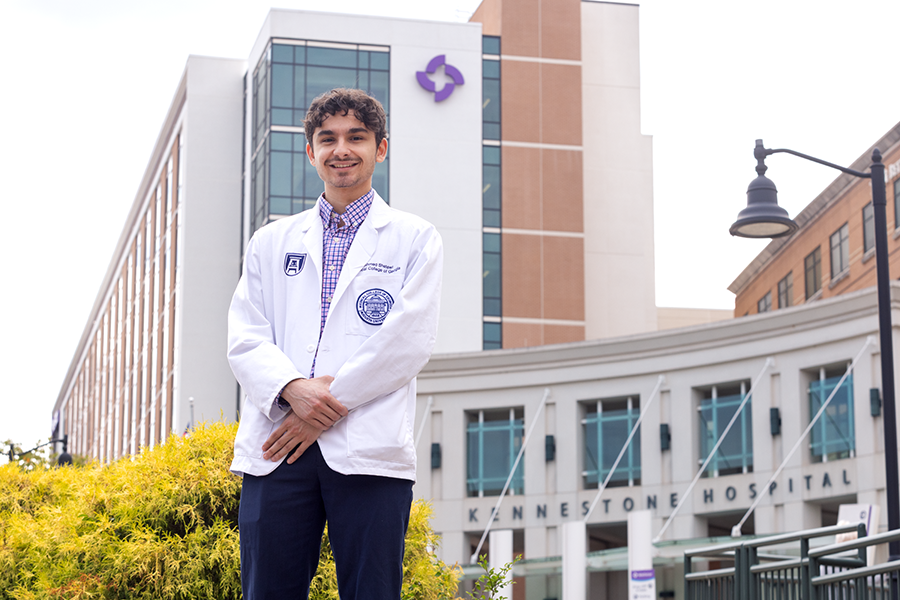 A medical college student wearing a lab coat stands in front of a hospital.