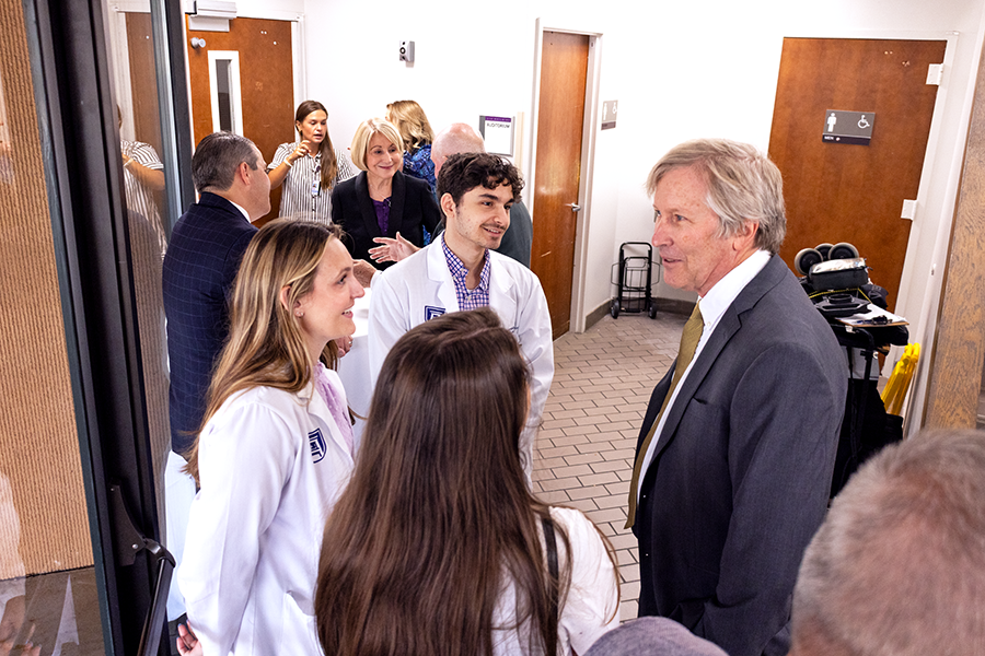 An older man and three medical students wearing lab coats stand in a loose circle and talk in the entryway to a hospital.