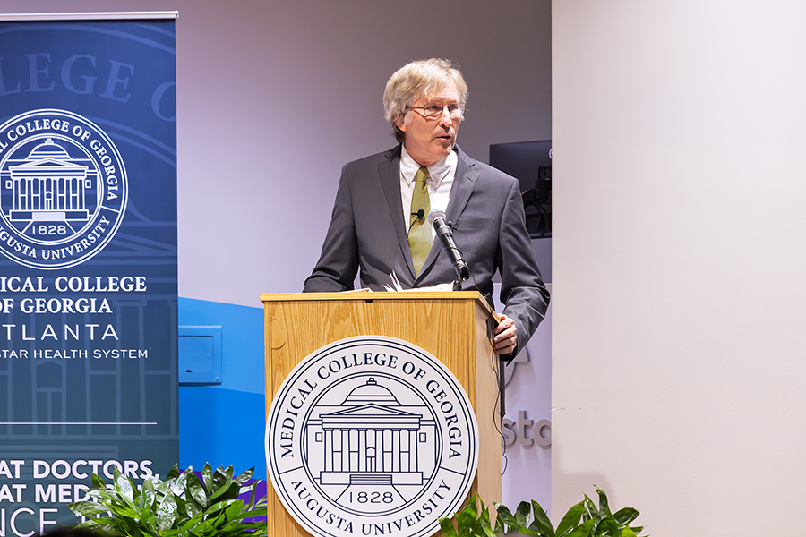 A man stands at a podium with a microphone and speaks to a large crowd in an auditorium. There is a seal for the Medical College of Georgia placed on the podium in front of him.