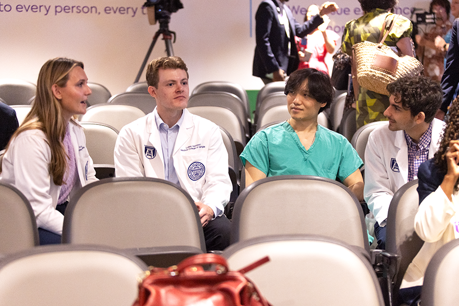 Four medical students, three wearing lab coats and one wearing scrubs, sit in a large auditorium.