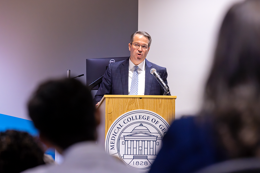 A man stands at a podium with a microphone and speaks to a large crowd in an auditorium. There is a seal for the Medical College of Georgia placed on the podium in front of him.
