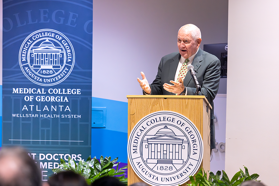 A man stands at a podium with a microphone and speaks to a large crowd in an auditorium. There is a seal for the Medical College of Georgia placed on the podium in front of him.