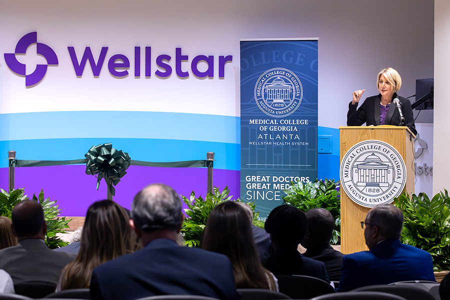 A woman stands at a podium with a microphone and speaks to a large crowd in an auditorium. There is a seal for the Medical College of Georgia placed on the podium in front of her.