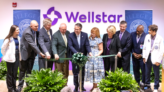 A group of 10 people take part in a ceremonial ribbon cutting ceremony on a stage in front of a larger group of people.