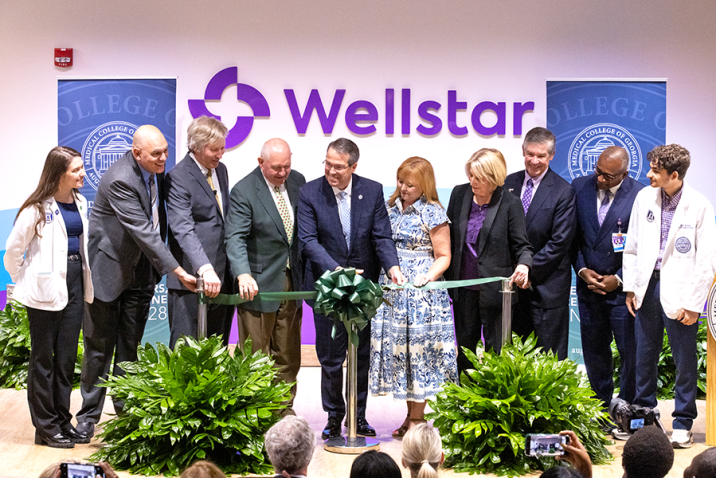 A group of 10 people take part in a ceremonial ribbon cutting ceremony on a stage in front of a larger group of people.