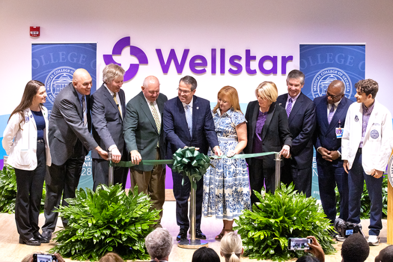 A group of 10 people take part in a ceremonial ribbon cutting ceremony on a stage in front of a larger group of people.