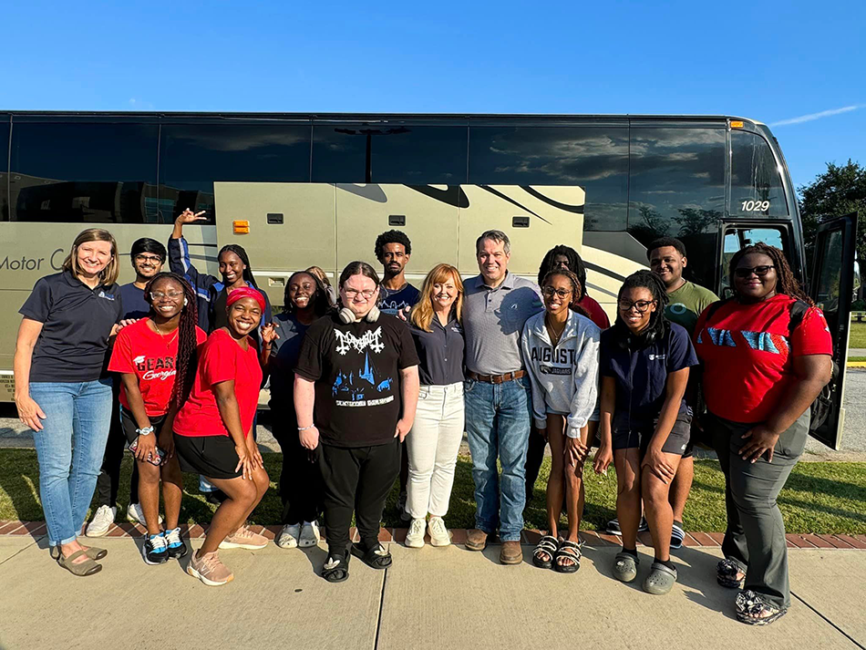 Young men and women pose with man and two women in front of large bus.