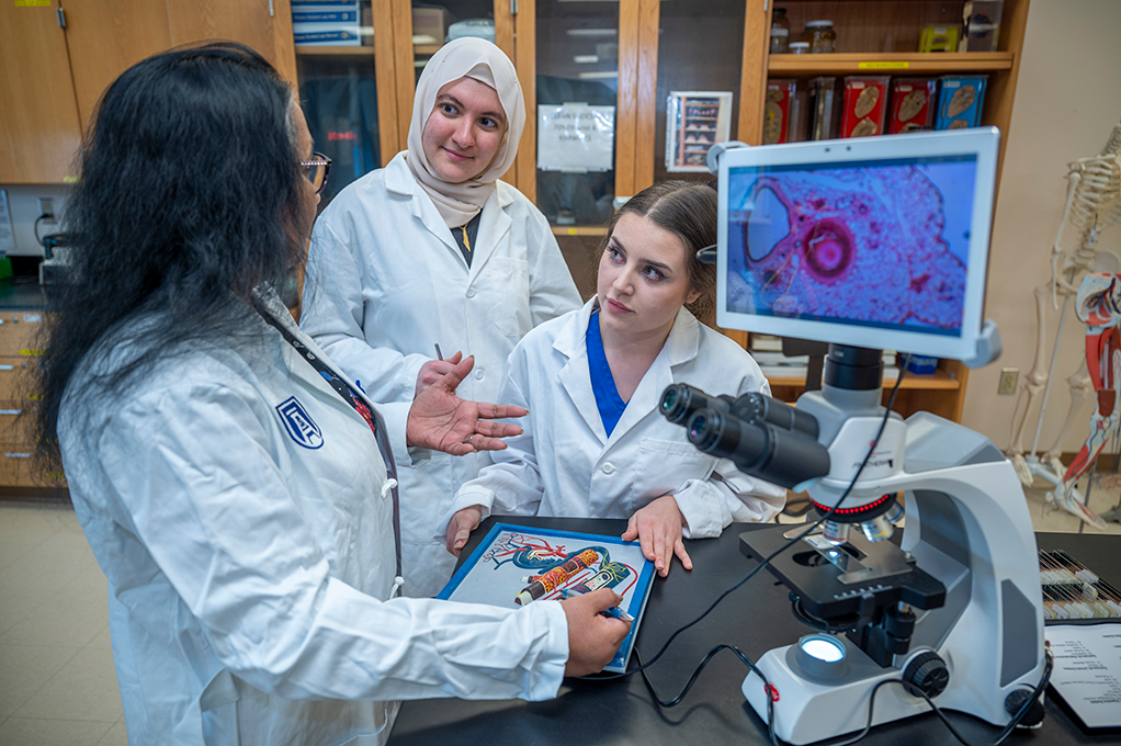 Three women in white coats talk over medical models and a telescope.