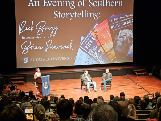 Two men sit in chairs on a stage in front of a large group of people gathered for a presentation. There is also a woman standing at a podium with a microphone off to the side talking about the event.