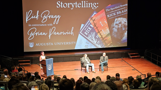 Two men sit in chairs on a stage in front of a large group of people gathered for a presentation. There is also a woman standing at a podium with a microphone off to the side talking about the event.