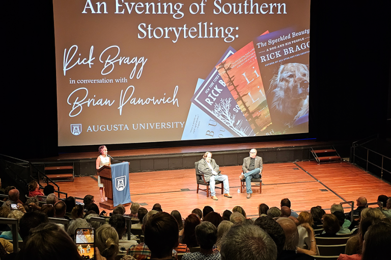 Two men sit in chairs on a stage in front of a large group of people gathered for a presentation. There is also a woman standing at a podium with a microphone off to the side talking about the event.
