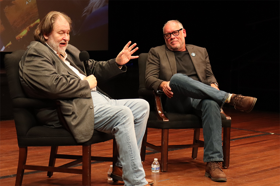 Two men sit in chairs on a stage in front of a large group of people gathered for a presentation. They are both holding microphones as one is telling a story to the crowd.