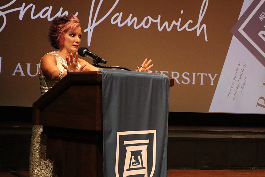 A woman stands at a podium with a microphone and introduces a program to a large group of people gathered in an auditorium.