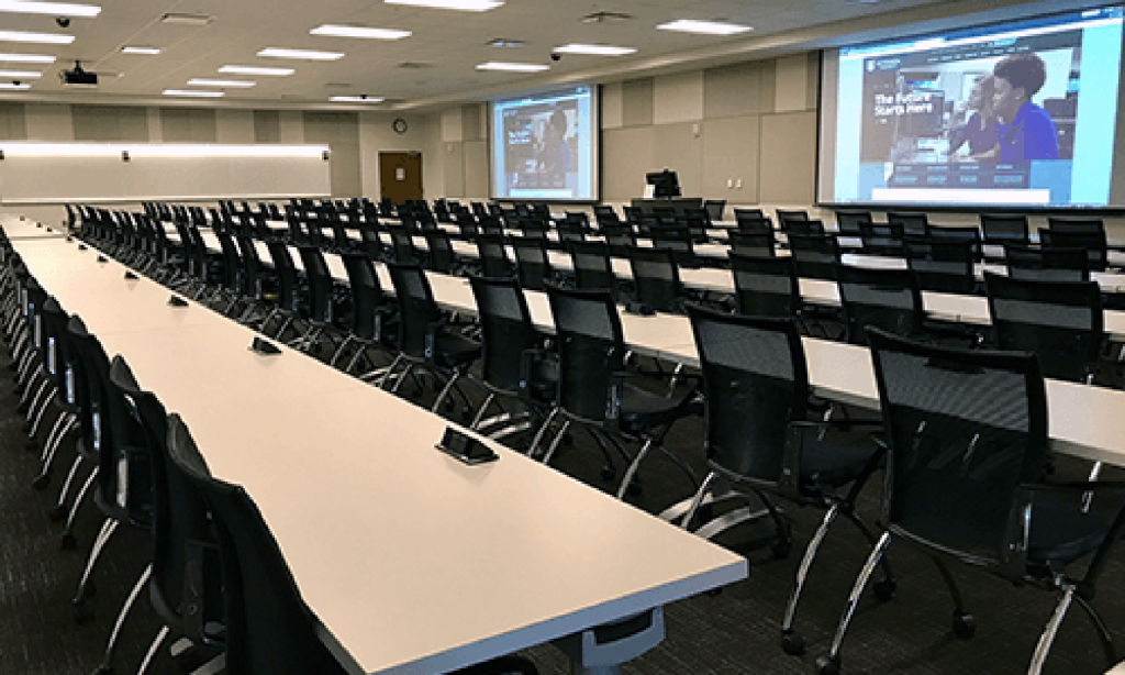A large classroom with long tables and chairs that are facing two projector screens.