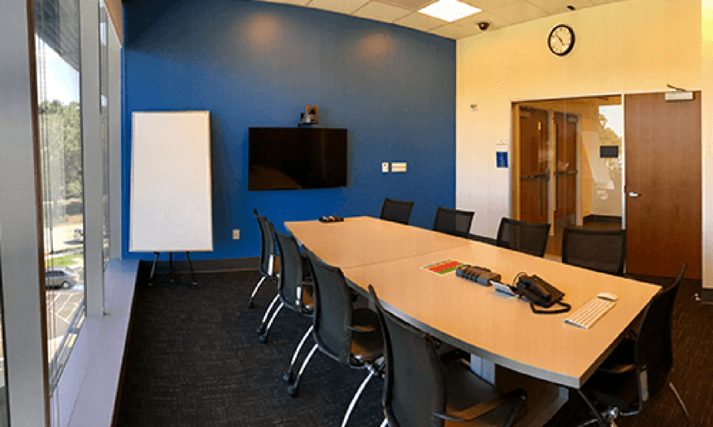 A conference room complete with a large table and chairs. A large screen is on the wall and a dry erase board beside it.