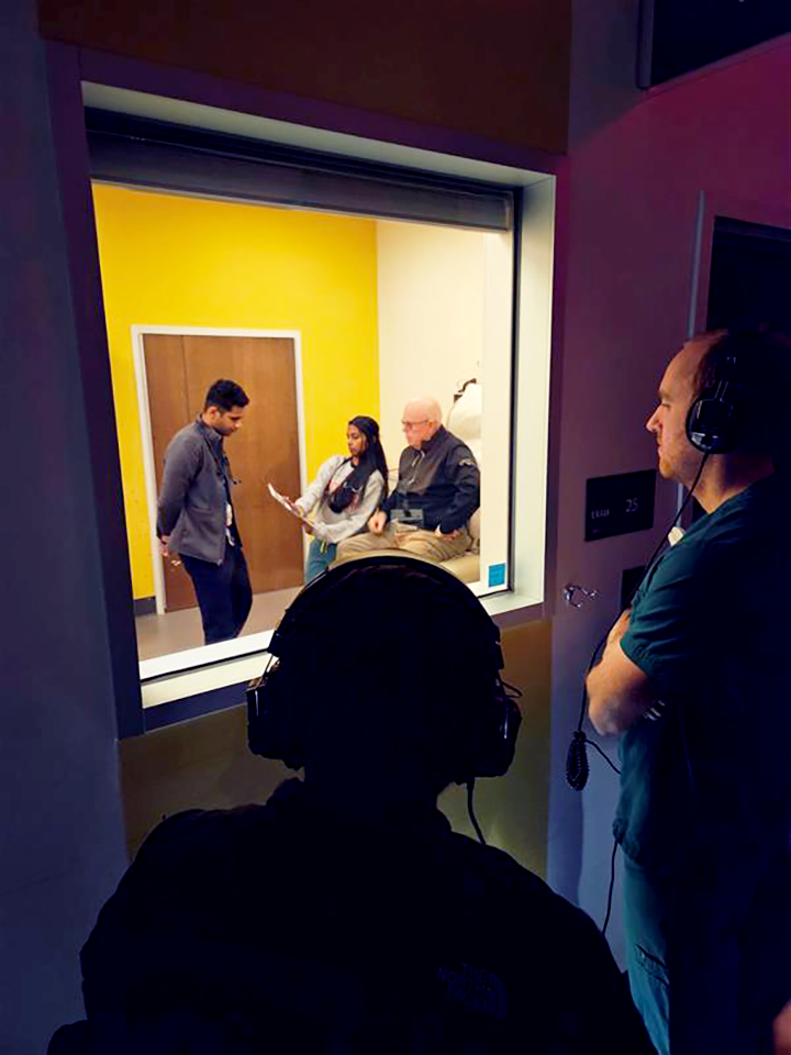 Two people wearing headphones observe a medical patient and two medical students throw a glass window.