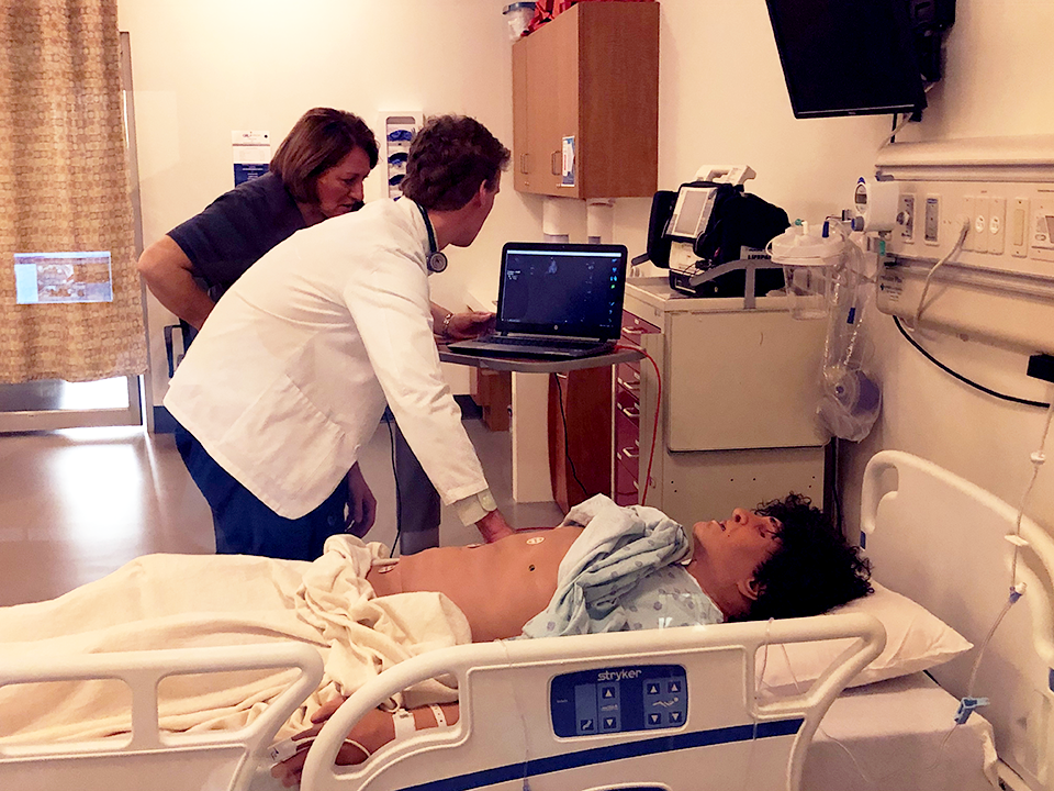 Two students practicing medicine stand over a medical manikin in a hospital bed in a simulated hospital room.