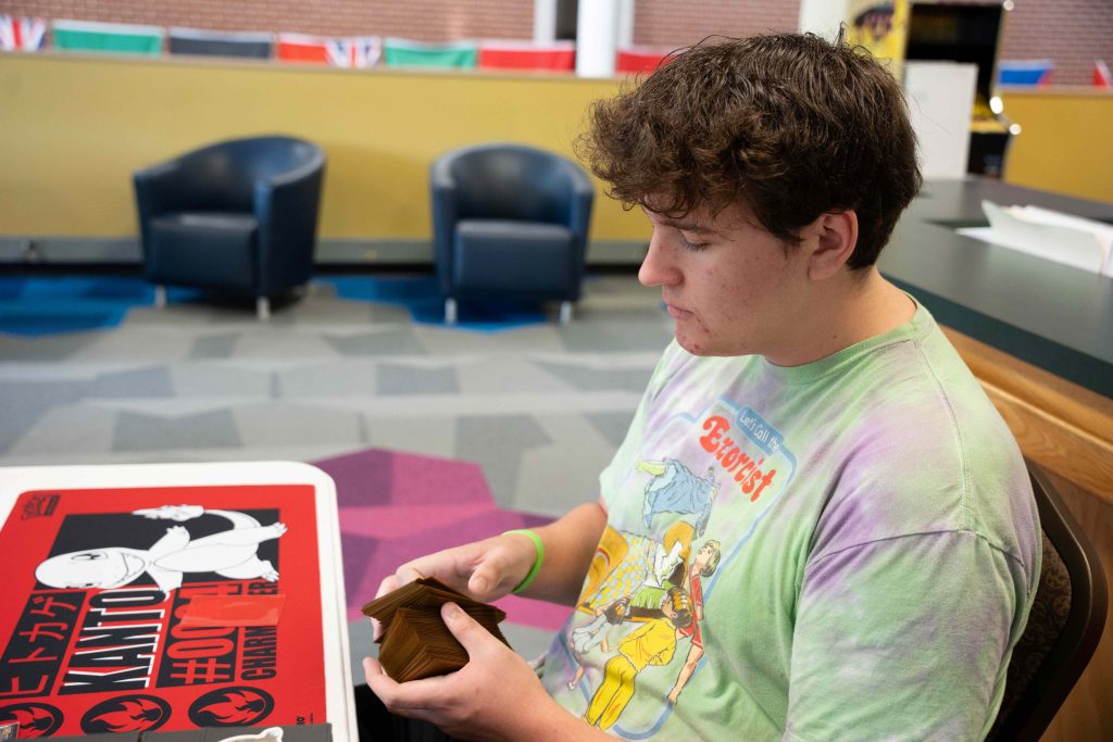 A college-aged male student sits at a table and is shuffling a deck of cards.