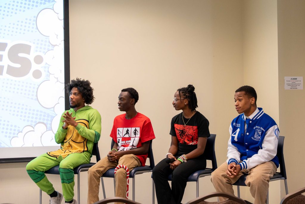A group of four male college students sit on tall chairs as part of a panel during a cosplay event.