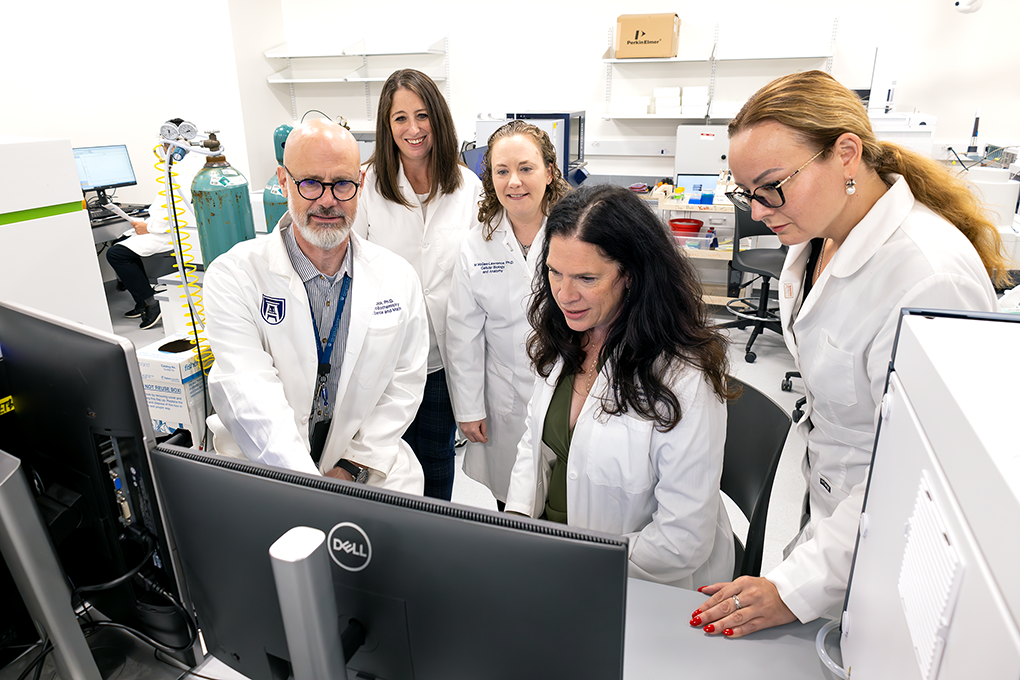Five university faculty researchers wearing lab coats huddle around a computer in a laboratory.
