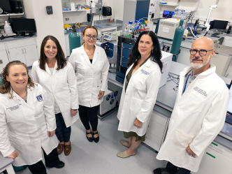 A group of university faculty researchers in lab coats stand in a laboratory and pose for the camera.