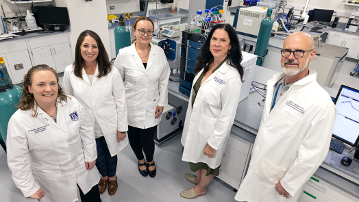 A group of university faculty researchers in lab coats stand in a laboratory and pose for the camera.