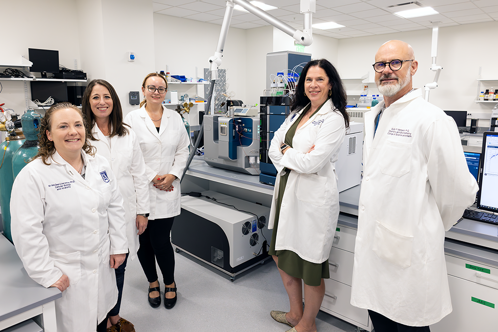 Five university faculty researchers wearing lab coats stand in a lab and smile at the camera.