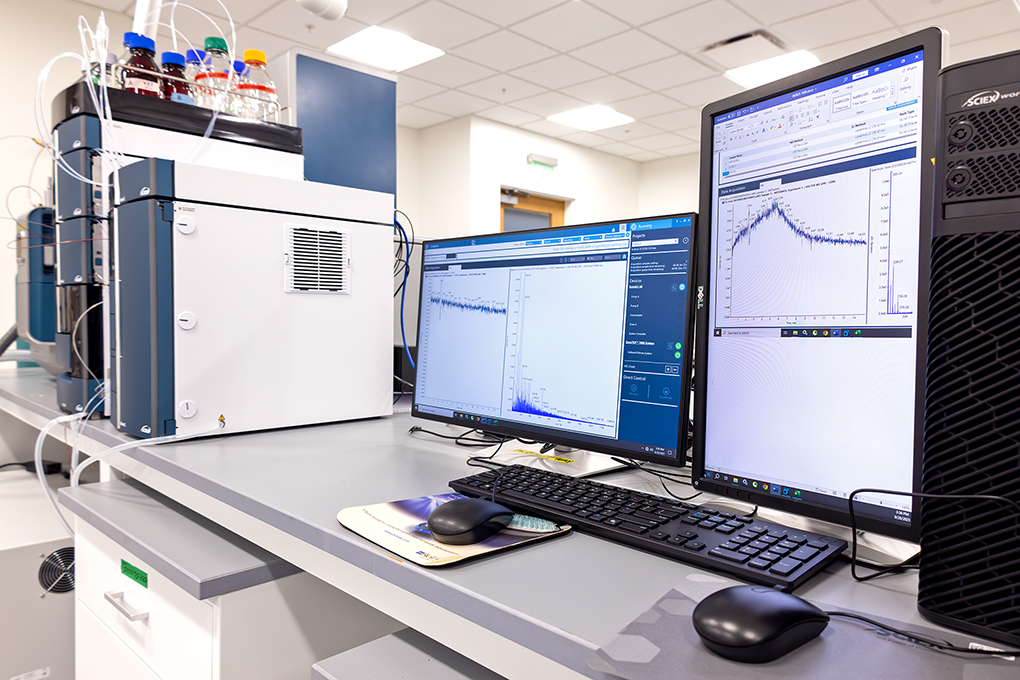 A horizontal and a vertical computer monitor sit beside a mass spectrometer on a table in a lab.