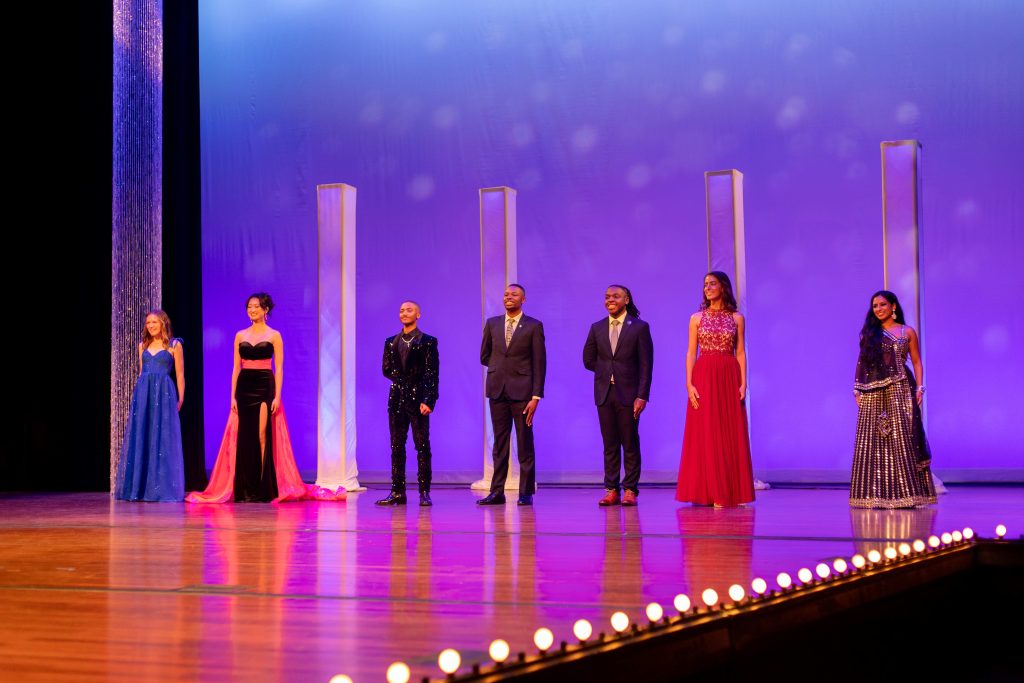 College-aged students dressed in evening wear stand in line on a stage.