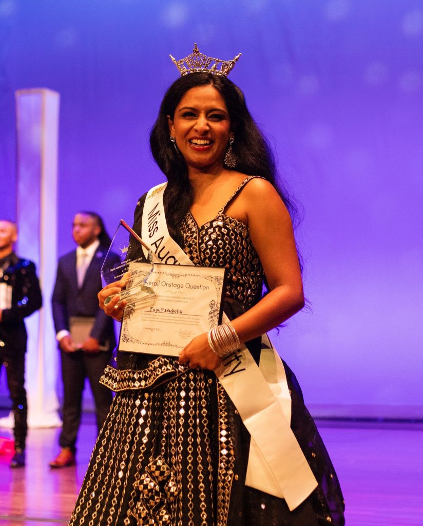 A female college student in a fancy ballgown celebrates being crowned Miss Augusta University. She is holding a certificate and wearing a crown and sash.