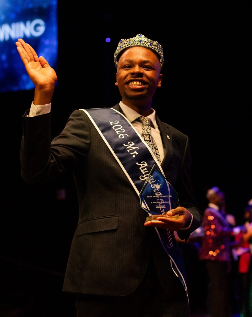 A man is standing on a stage wearing a crown and sash, while holding a trophy and plaque.