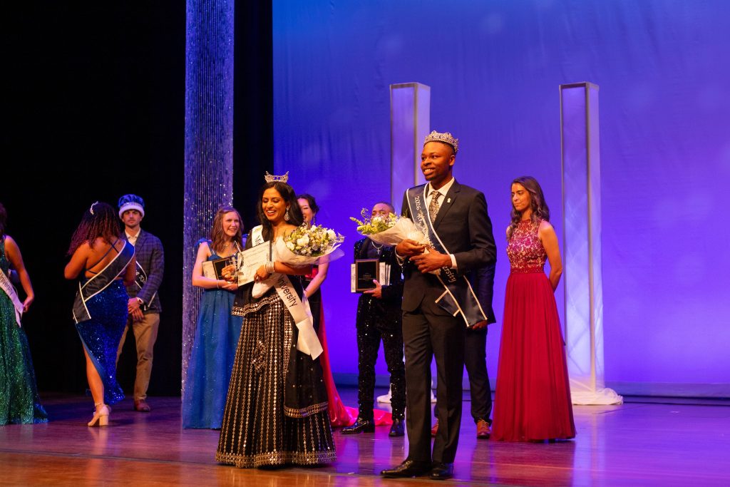 A man and woman wearing evening wear stand on a stage while holding a trophy, plaque and bouquet of flowers.
