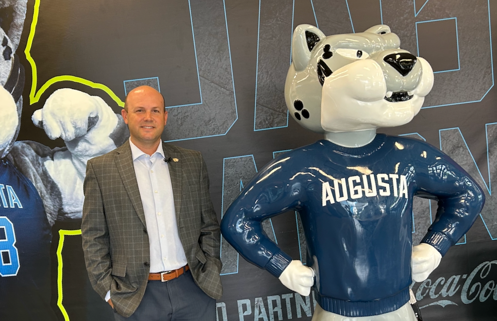 A man stands next to a statue that depicts a jaguar wearing a navy sweater with "Augusta" written across it in white.
